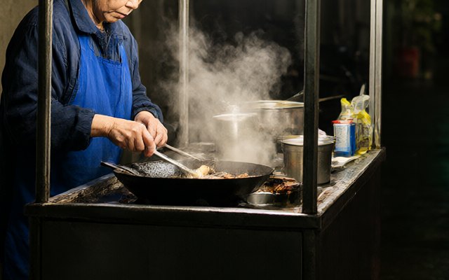 Close-up of the grandmother's hands in the second street render: her right hand holds tongs over the wok, and an extra spoon lies in the wok by her left hand with a handle that doesn't lead anywhere